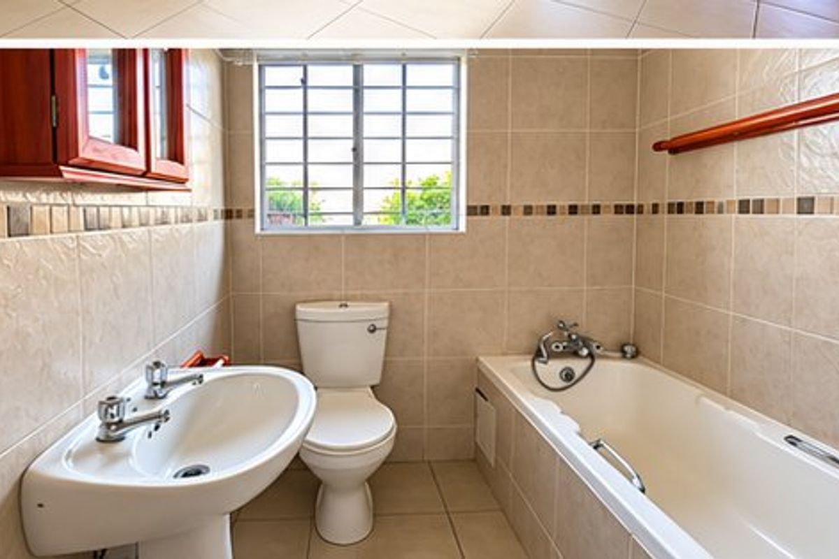 A functional bathroom featuring a pedestal basin, toilet, and bathtub with neutral wall and floor tiling, complemented by a window with security bars.