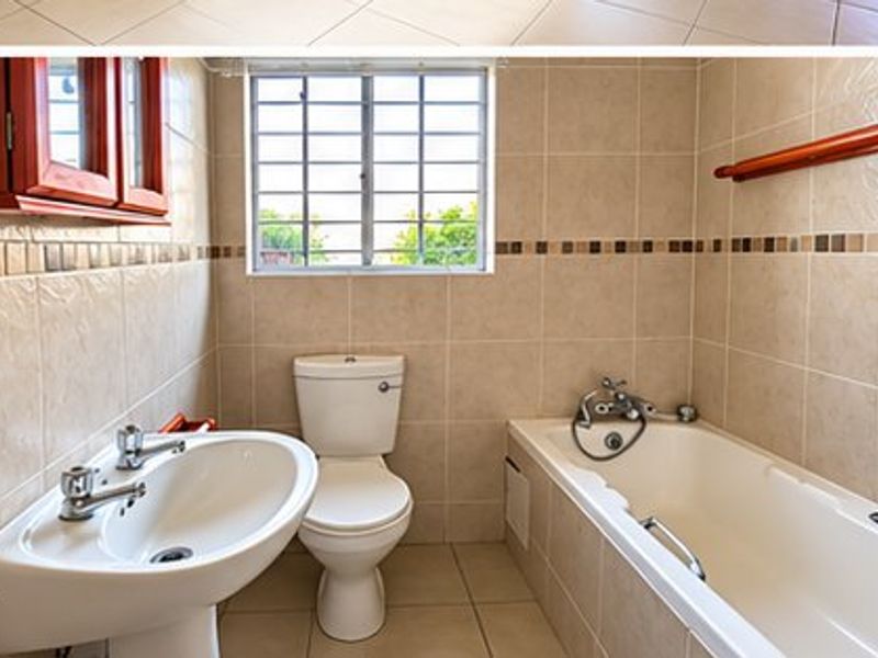 A functional bathroom featuring a pedestal basin, toilet, and bathtub with neutral wall and floor tiling, complemented by a window with security bars.
