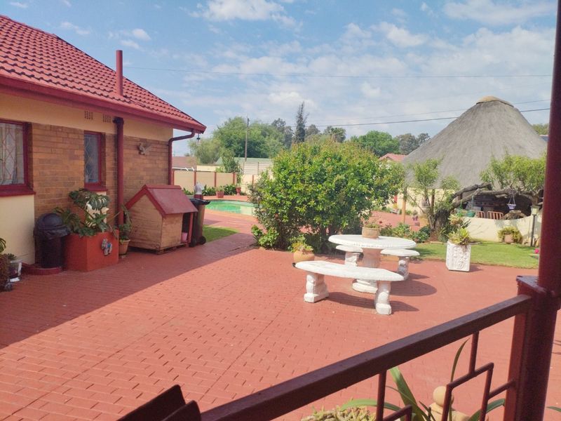 Rear exterior view featuring a paved patio area, swimming pool, and a traditional thatched lapa structure in the garden.
