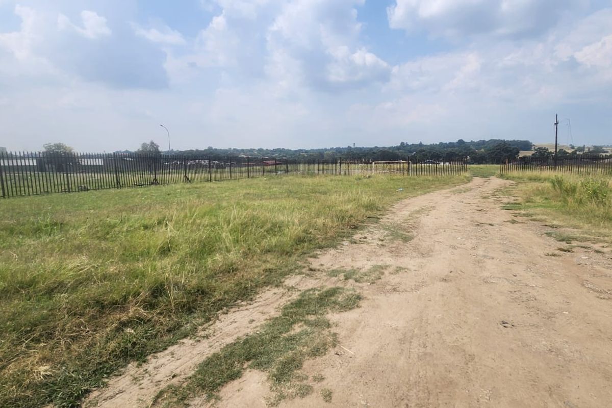 A large, undeveloped industrial plot featuring an unpaved access road and secure steel palisade perimeter fencing.