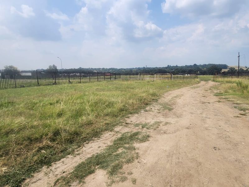 A large, undeveloped industrial plot featuring an unpaved access road and secure steel palisade perimeter fencing.