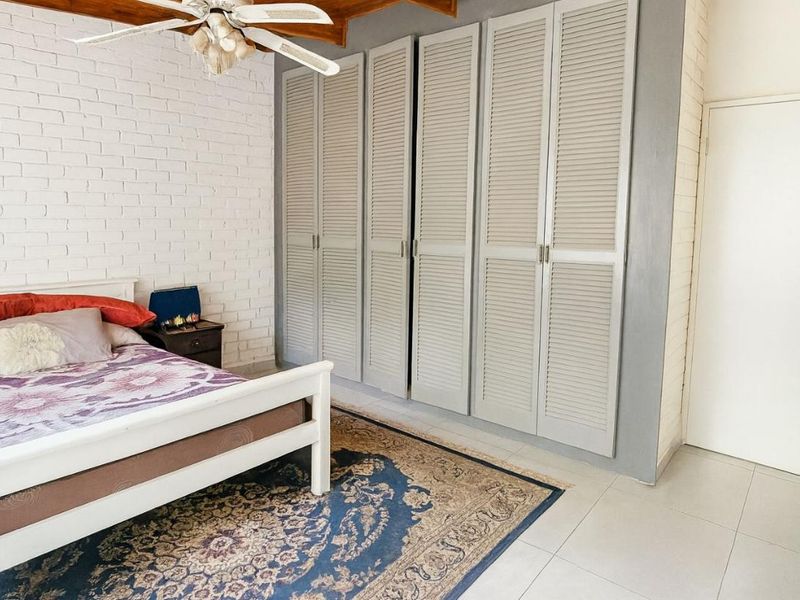 Bedroom featuring built-in louvered cupboards, exposed white brick wall, tiled flooring, and a ceiling fan.
