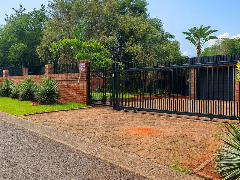 Front view of the property featuring a secure metal gate, brick boundary wall, and a well-maintained paved driveway with landscaped garden elements.