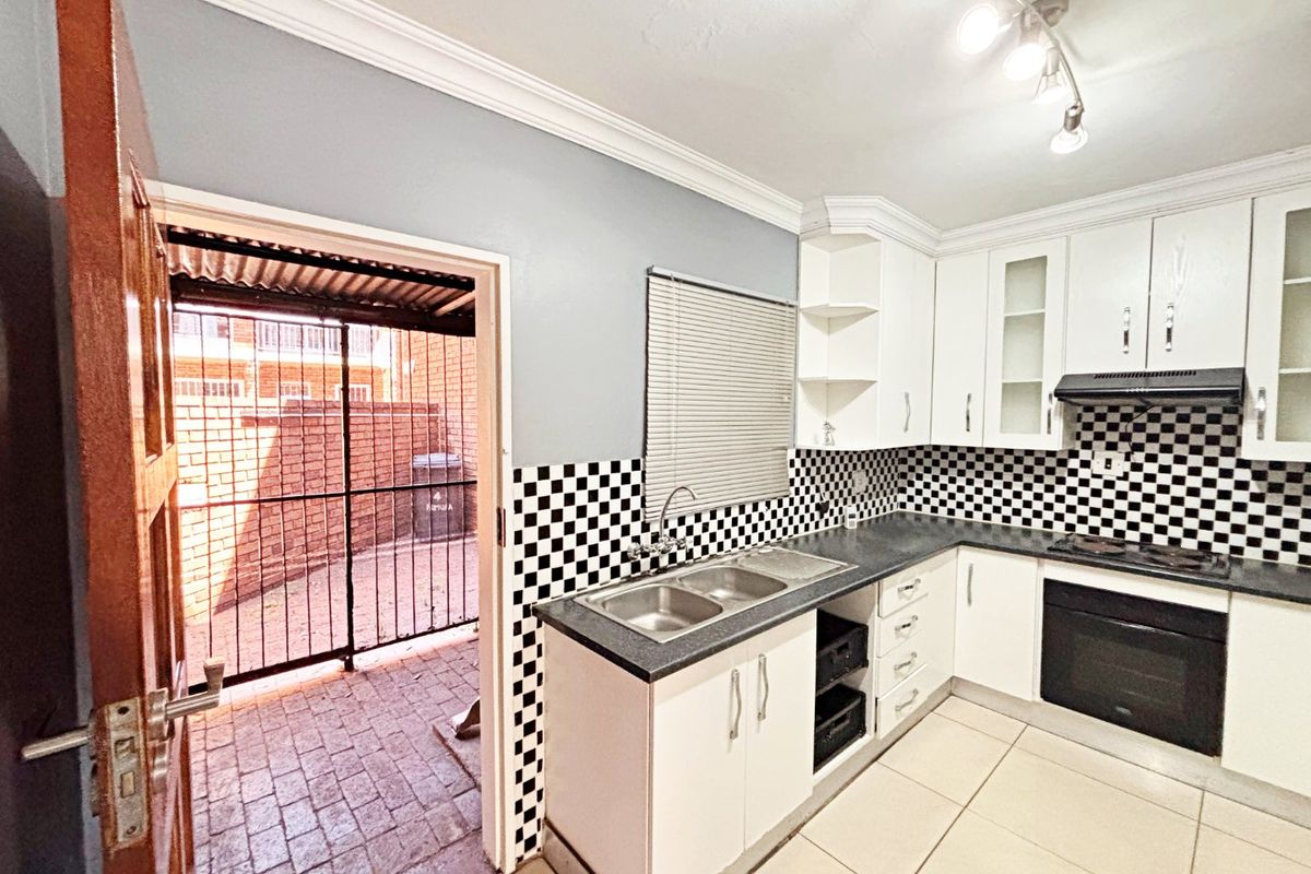 A functional kitchen featuring white cabinetry, a checkerboard tiled splashback, built-in oven and hob, and direct access to an outdoor paved area.