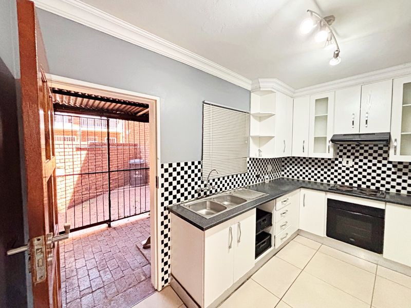 A functional kitchen featuring white cabinetry, a checkerboard tiled splashback, built-in oven and hob, and direct access to an outdoor paved area.