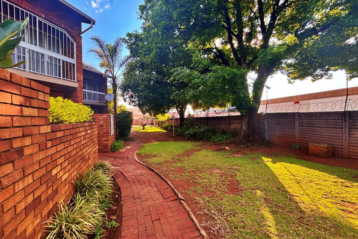 A well-maintained rear garden area featuring a brick paved pathway, mature trees, and secure perimeter walling with electric fencing.