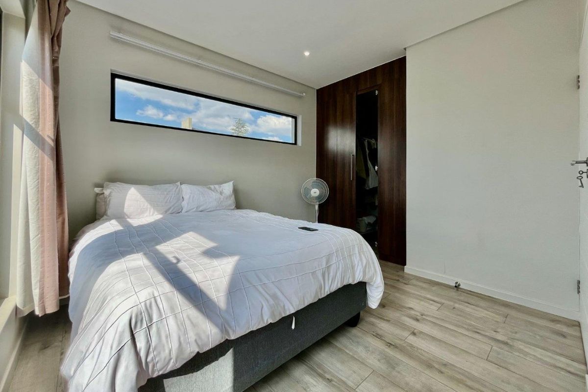 A modern bedroom featuring laminate flooring, built-in dark wood cupboards, and a horizontal clerestory window providing natural light.
