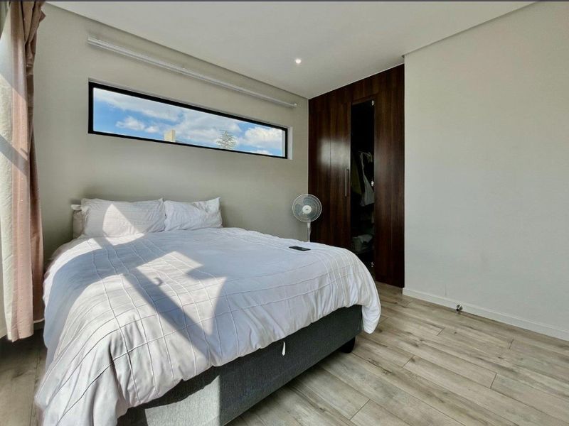 A modern bedroom featuring laminate flooring, built-in dark wood cupboards, and a horizontal clerestory window providing natural light.