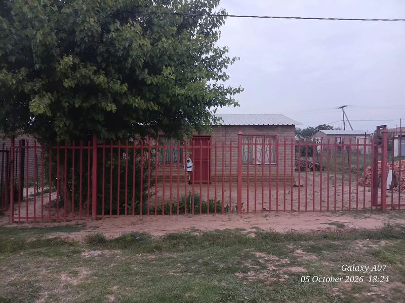 Front view of a single-story brick house featuring a metal security fence, security bars on windows, and a corrugated metal roof.
