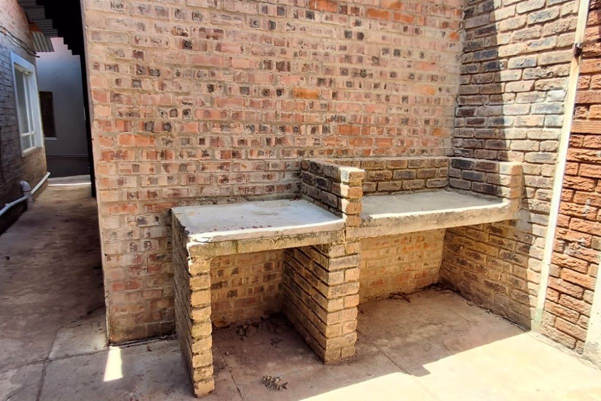 An outdoor rear area featuring a built-in brick braai with concrete countertops, set against exposed brick walls and paved flooring.