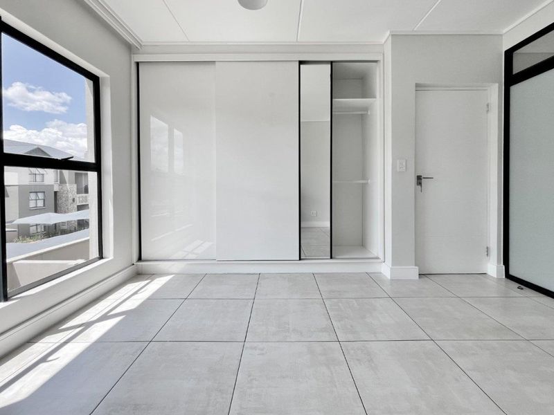 A bright, modern bedroom featuring large windows, neutral tiled flooring, and a sleek built-in sliding wardrobe with integrated shelving.