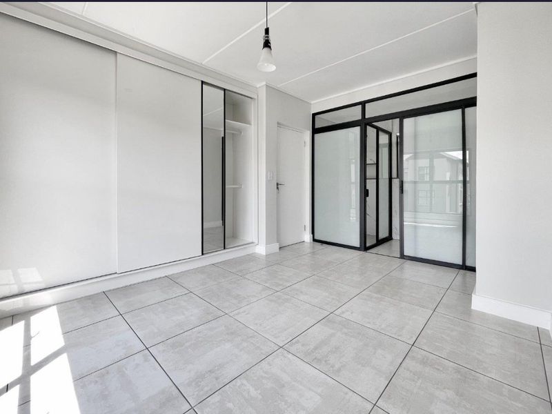 A modern, bright bedroom featuring large-format grey floor tiles, built-in sliding wardrobes, and a contemporary black-framed glass partition leading to an adjacent space.