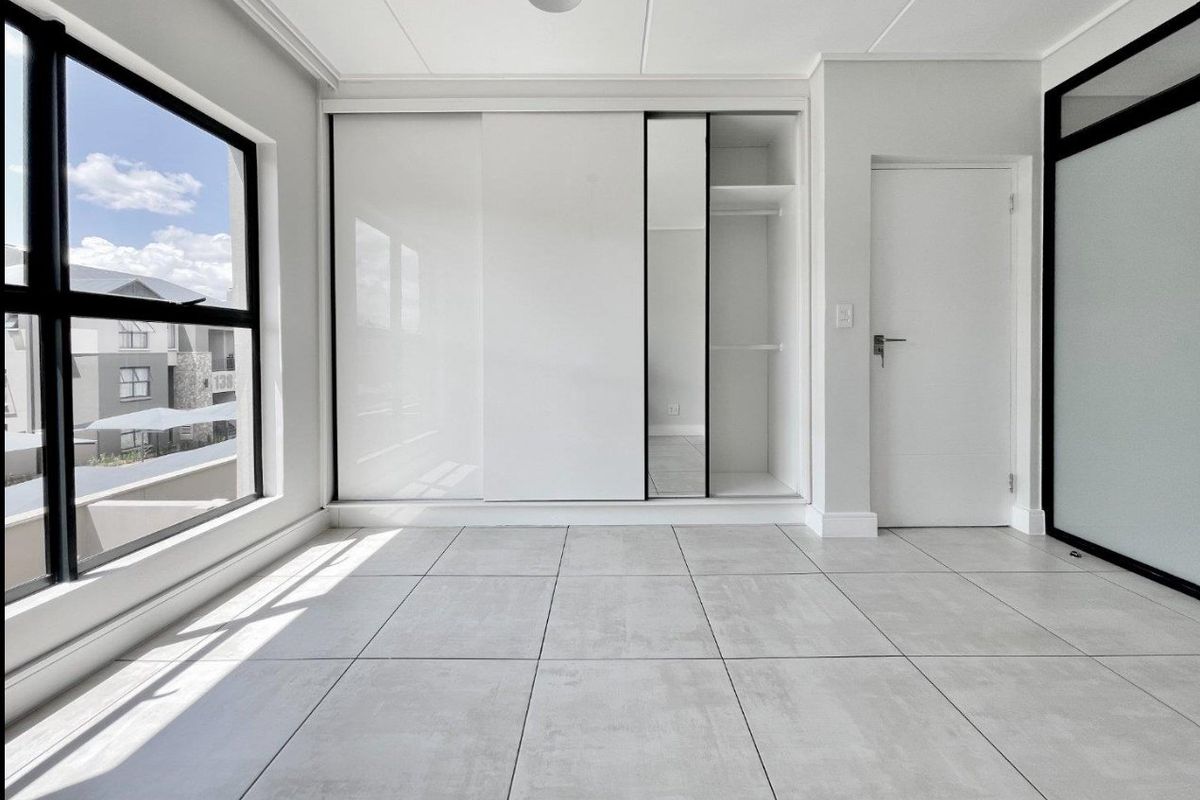 A bright, modern bedroom featuring large windows, neutral tiled flooring, and a sleek built-in sliding wardrobe with integrated shelving.