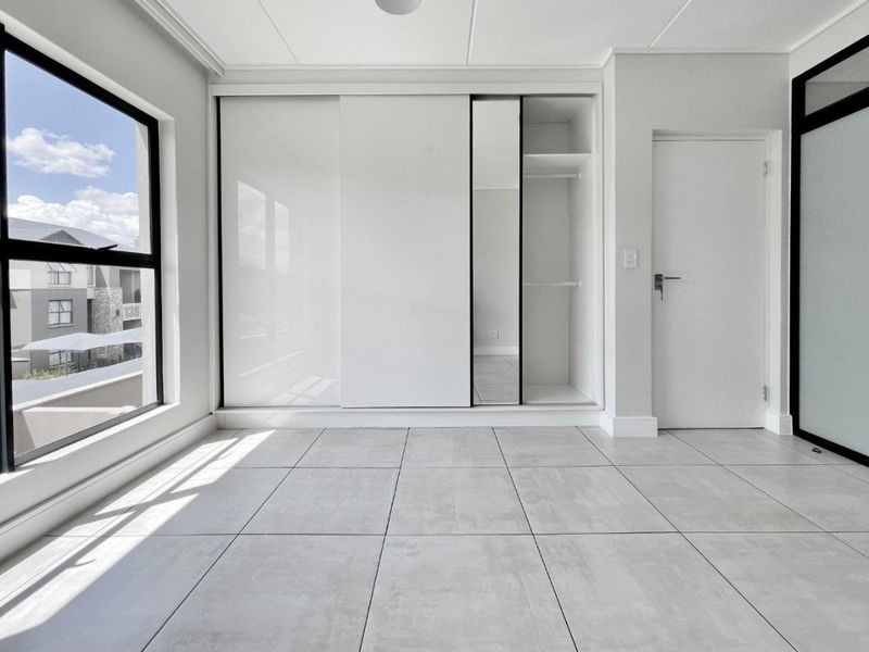 A bright, modern bedroom featuring large windows, neutral tiled flooring, and a sleek built-in sliding wardrobe with integrated shelving.