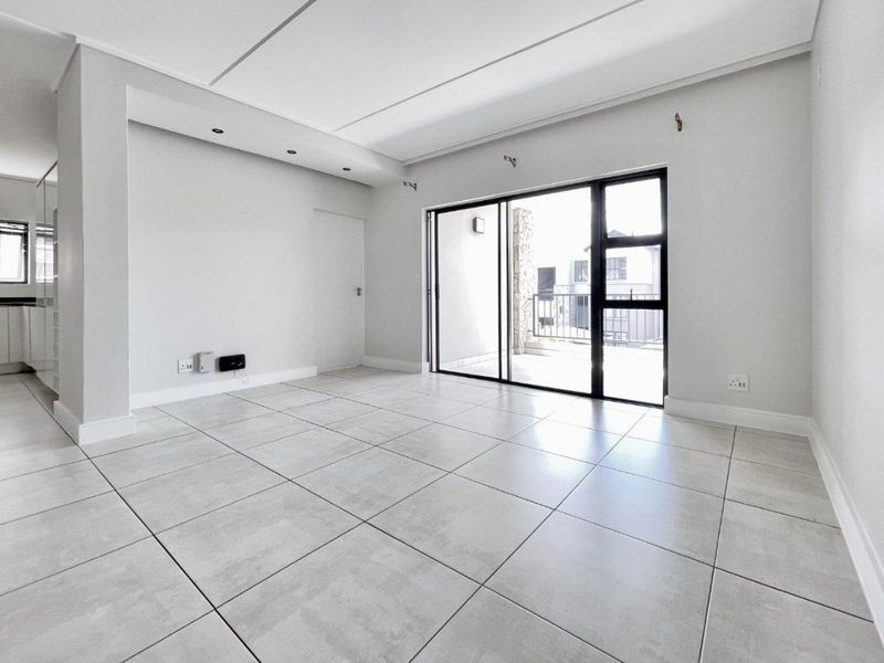A bright, empty open-plan living area featuring large format floor tiles, recessed ceiling lighting, and a sliding glass door leading to an outdoor balcony.