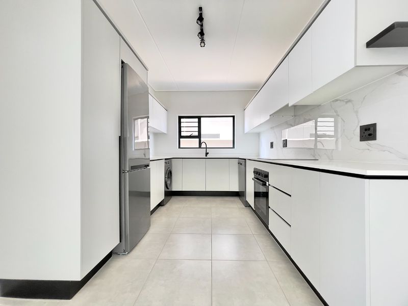 Modern galley-style kitchen featuring sleek white cabinetry, stone countertops, marble-look splashback, and integrated appliances including an oven, dishwasher, and washing machine.