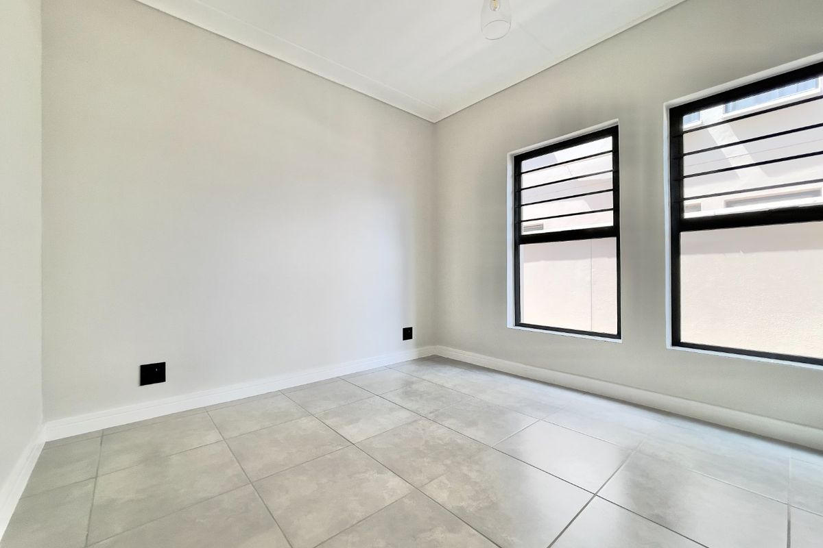 A bright, empty bedroom featuring neutral tiled flooring, modern black-framed windows with security bars, and clean, minimalist finishes.