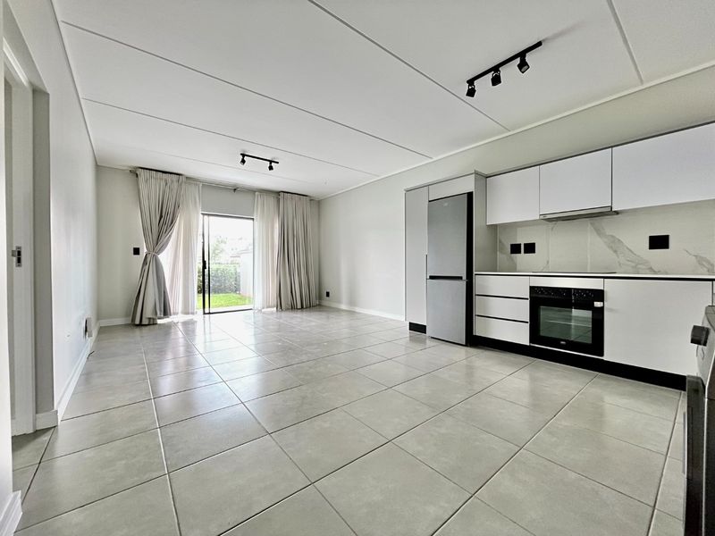 Modern open-plan living area featuring sleek white kitchen cabinetry, built-in oven, tiled floors, and direct access to a private outdoor space through sliding doors.