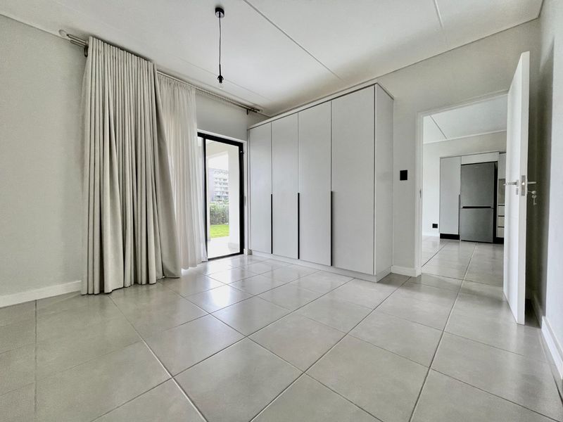 A bright, empty bedroom featuring neutral tiled flooring, modern built-in cupboards, and a sliding glass door providing access to the exterior.