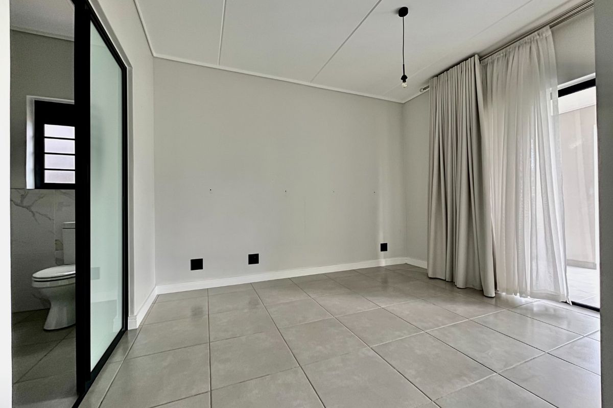 A bright, empty bedroom featuring large format floor tiles, a modern black-framed glass partition leading to an en-suite bathroom, and access to a patio.