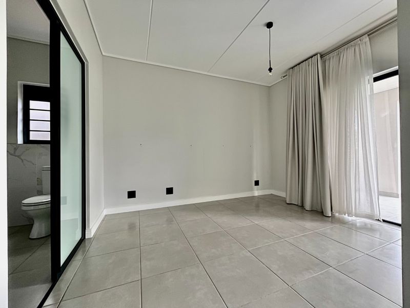 A bright, empty bedroom featuring large format floor tiles, a modern black-framed glass partition leading to an en-suite bathroom, and access to a patio.