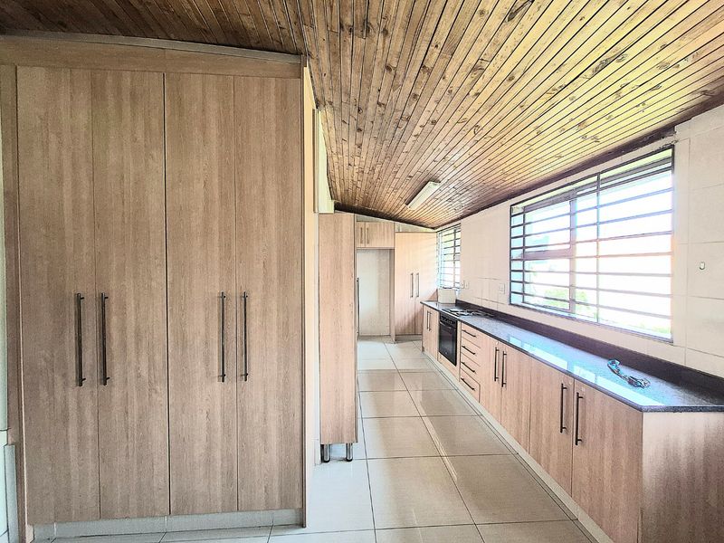 A spacious, empty kitchen featuring light wood-finish cabinetry, dark countertops, tiled flooring, and a distinctive timber-clad ceiling with integrated oven and hob.