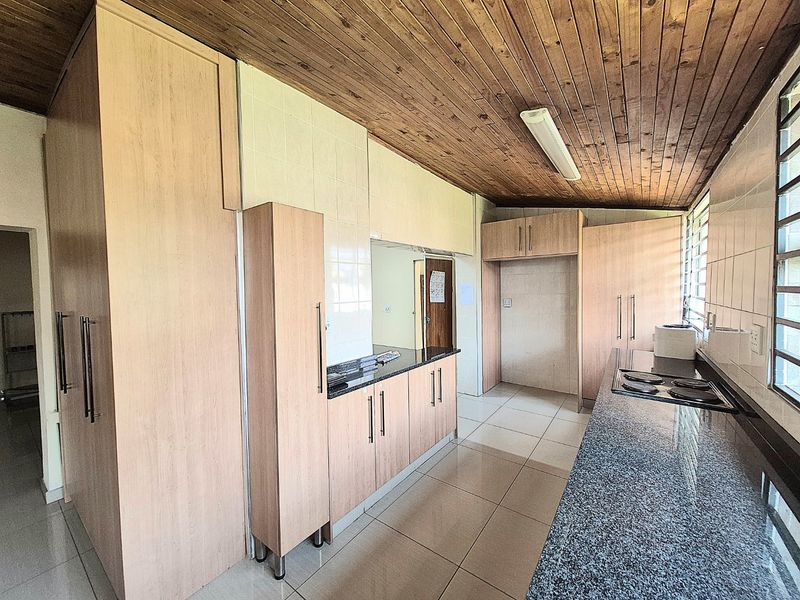A functional kitchen featuring light wood cabinetry, granite countertops, an integrated electric hob, tiled flooring, and a distinctive timber-paneled ceiling.