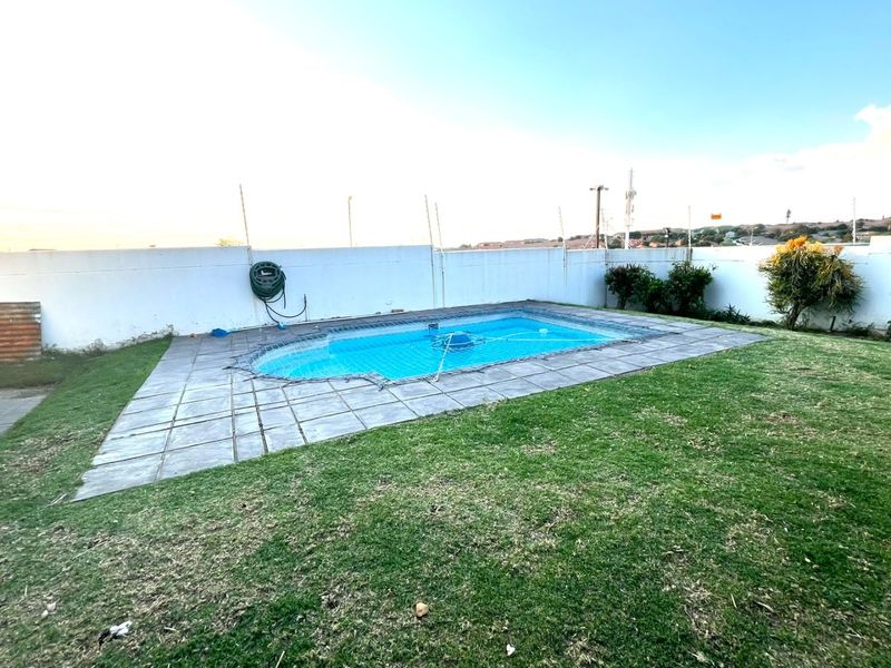Outdoor pool area featuring a swimming pool with a safety net, surrounded by a paved deck and a grass lawn with perimeter boundary walls.