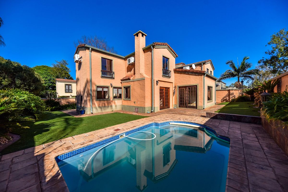 Rear view of a two-story house featuring a swimming pool, paved patio area, artificial lawn, and a prominent chimney stack.