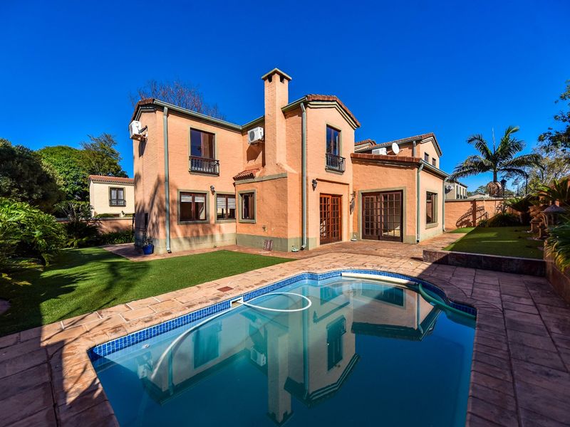 Rear view of a two-story house featuring a swimming pool, paved patio area, artificial lawn, and a prominent chimney stack.