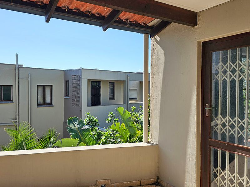 A covered balcony featuring tiled flooring, a security gate, and timber roof beams, offering an open view of the surrounding apartment complex.