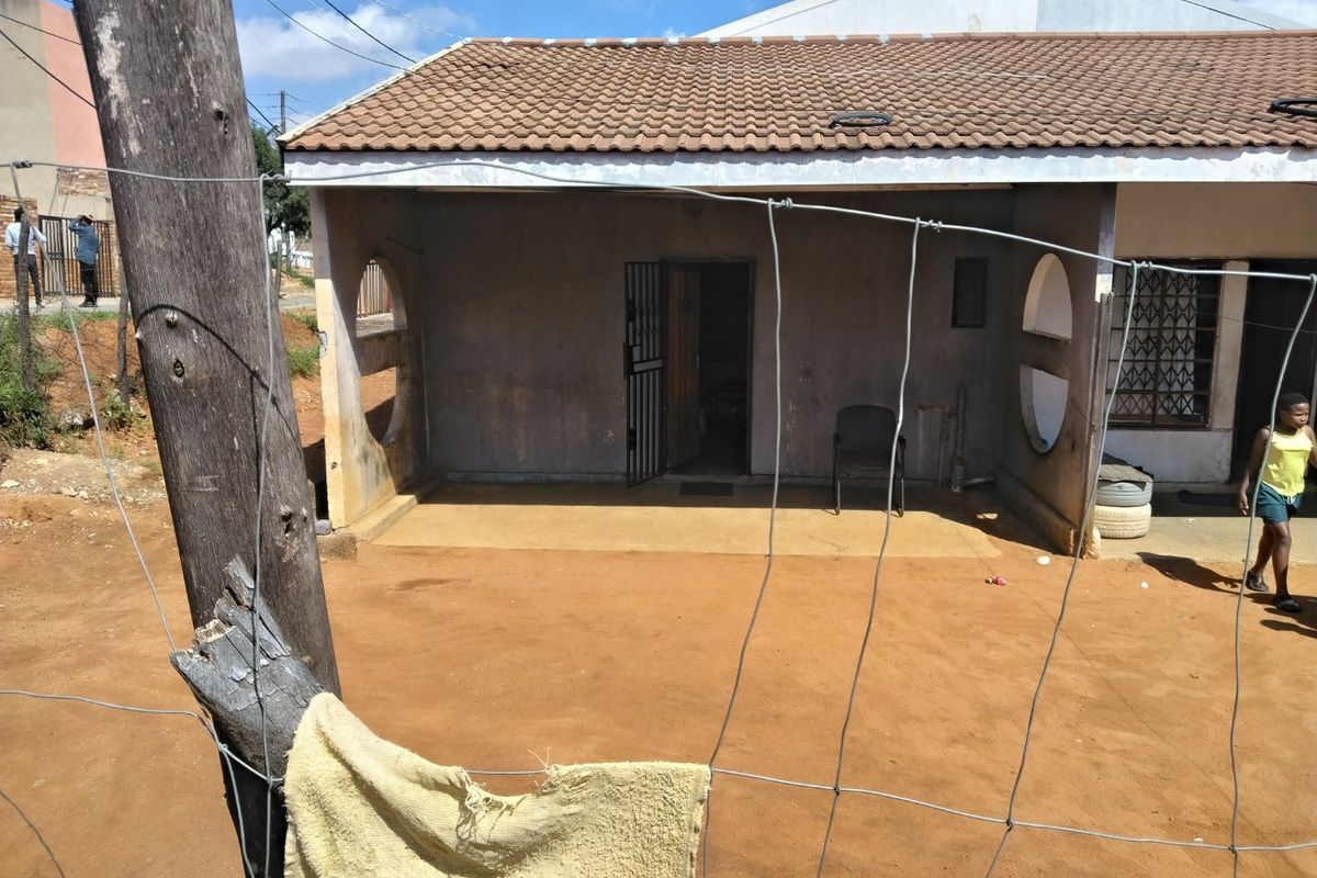 Rear view of the house featuring a covered patio area with security gate and tiled roof, set against a dirt yard.