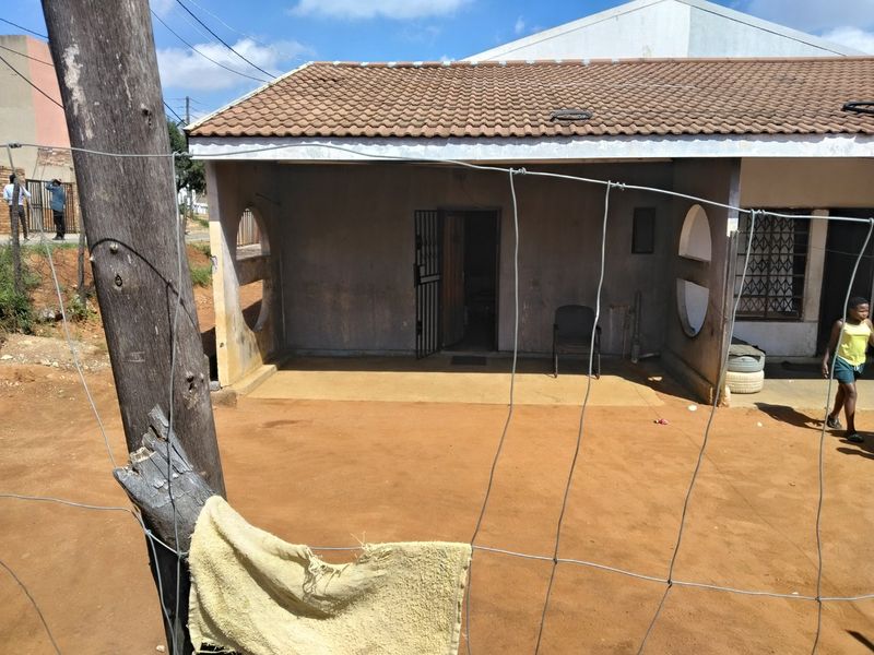Rear view of the house featuring a covered patio area with security gate and tiled roof, set against a dirt yard.