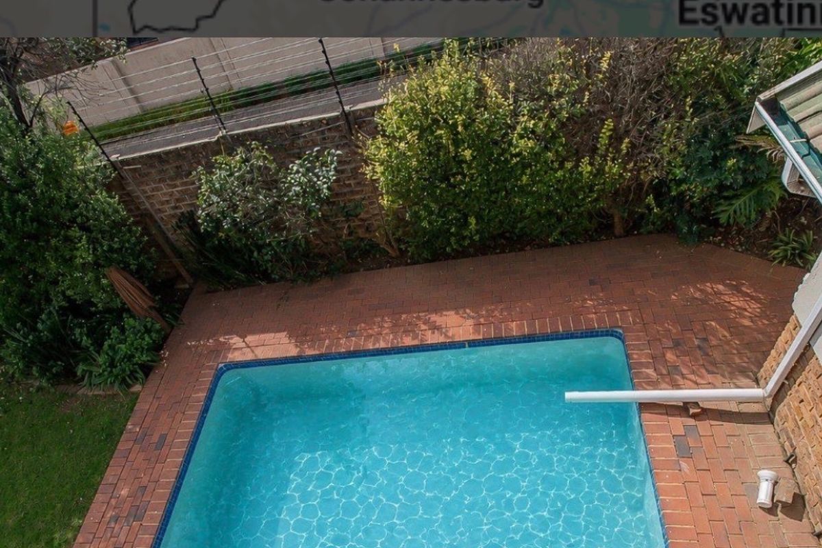 An elevated view of a private swimming pool area featuring brick paving, surrounding garden landscaping, and a secure boundary wall with electric fencing.