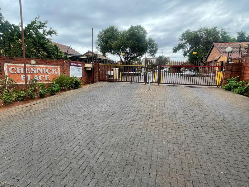 The entrance to Chesnick Place townhouse complex featuring a paved driveway, brick boundary wall, and secure automated gate access.