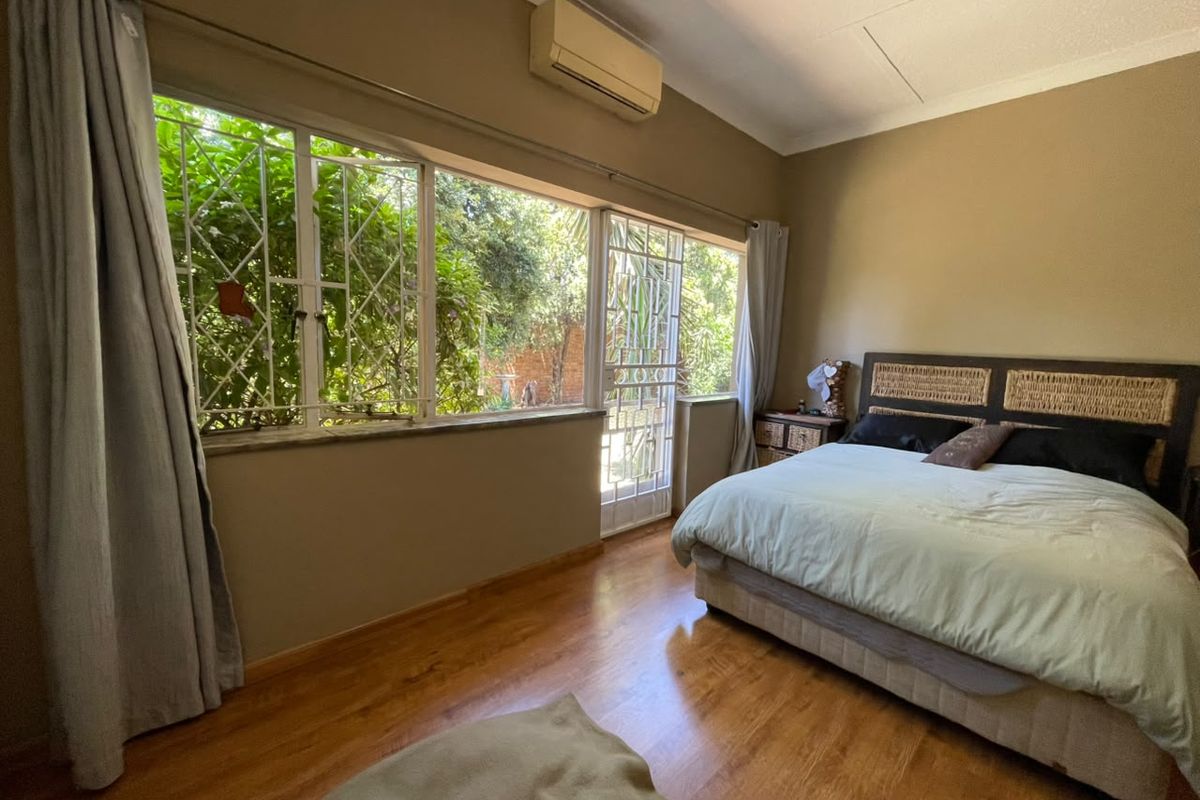 A bedroom featuring laminate flooring, a split-system air conditioner, and large windows with security bars, offering natural light and garden views.