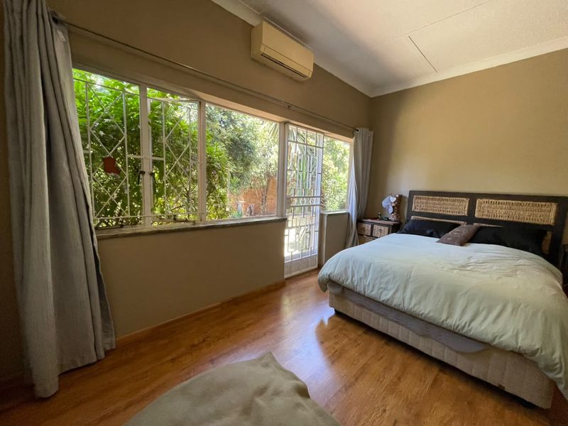 A bedroom featuring laminate flooring, a split-system air conditioner, and large windows with security bars, offering natural light and garden views.