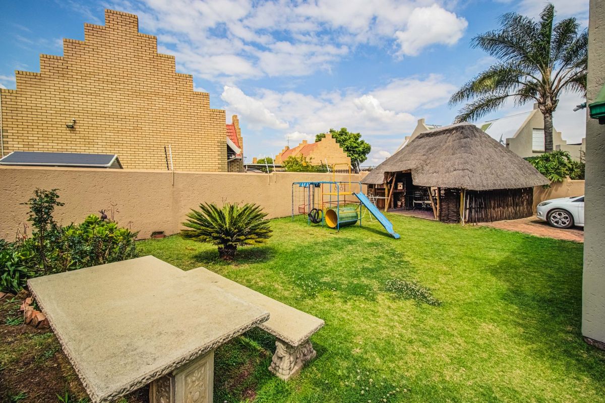 A spacious rear garden featuring a traditional thatched lapa, a concrete garden bench, and a well-maintained lawn area.