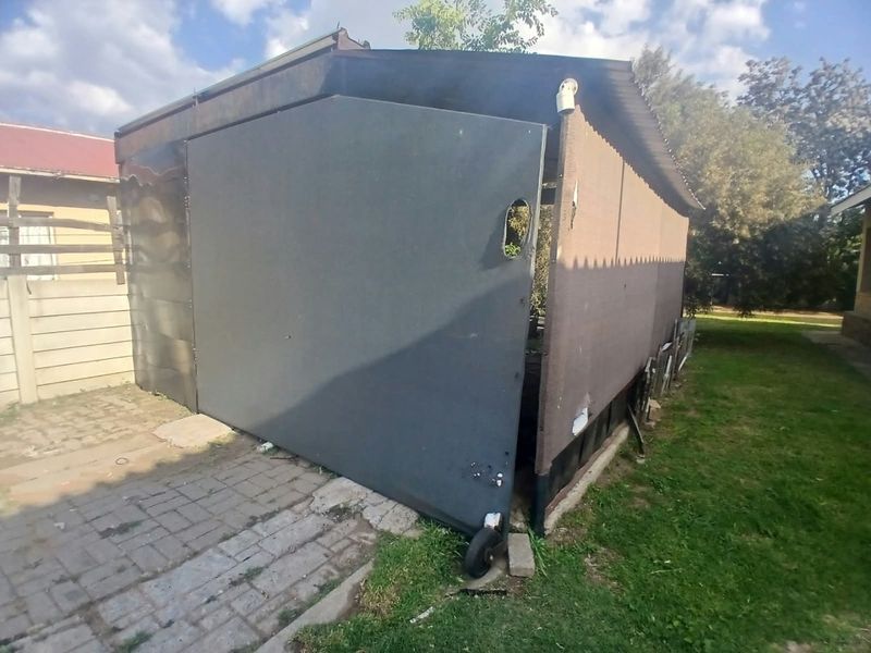 A rear exterior view showing a damaged sliding gate structure with corrugated metal roofing and adjacent paved area in a garden setting.
