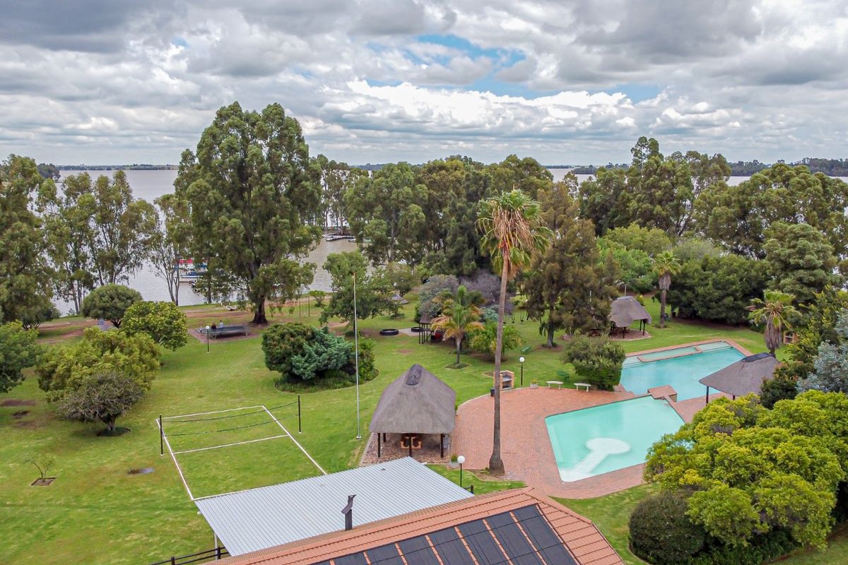 Aerial view of a spacious property featuring a large swimming pool, thatched lapa, volleyball court, and expansive garden overlooking the water.