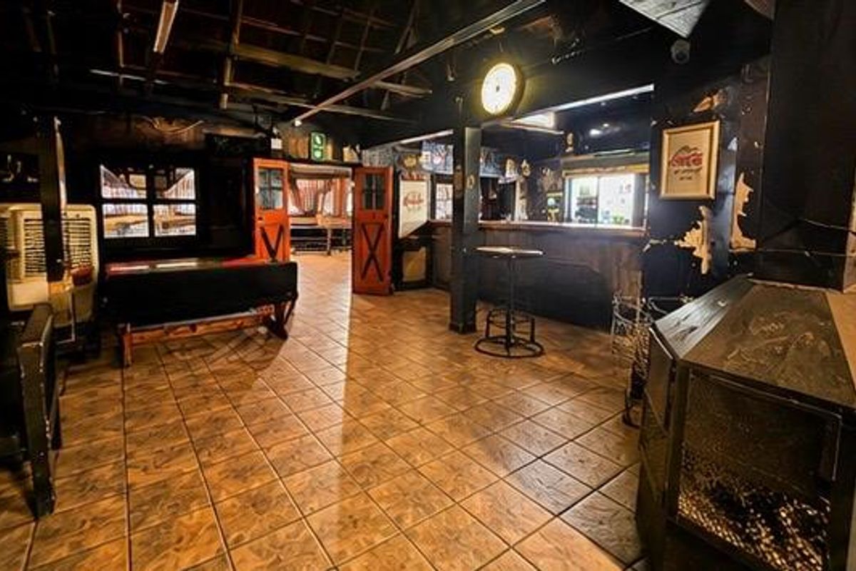 A rustic commercial bar area featuring tiled flooring, exposed ceiling beams, a built-in bar counter, and a central fireplace, requiring cosmetic updates to wall finishes.