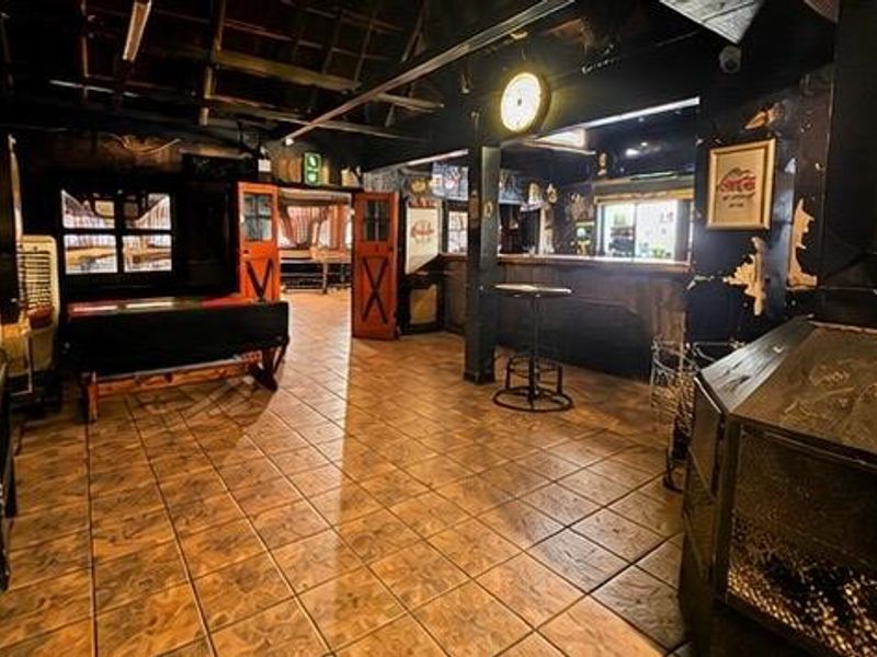 A rustic commercial bar area featuring tiled flooring, exposed ceiling beams, a built-in bar counter, and a central fireplace, requiring cosmetic updates to wall finishes.