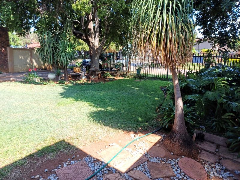 A well-maintained residential garden featuring a lush lawn, mature trees, and paved stepping stones enclosed by metal perimeter fencing.
