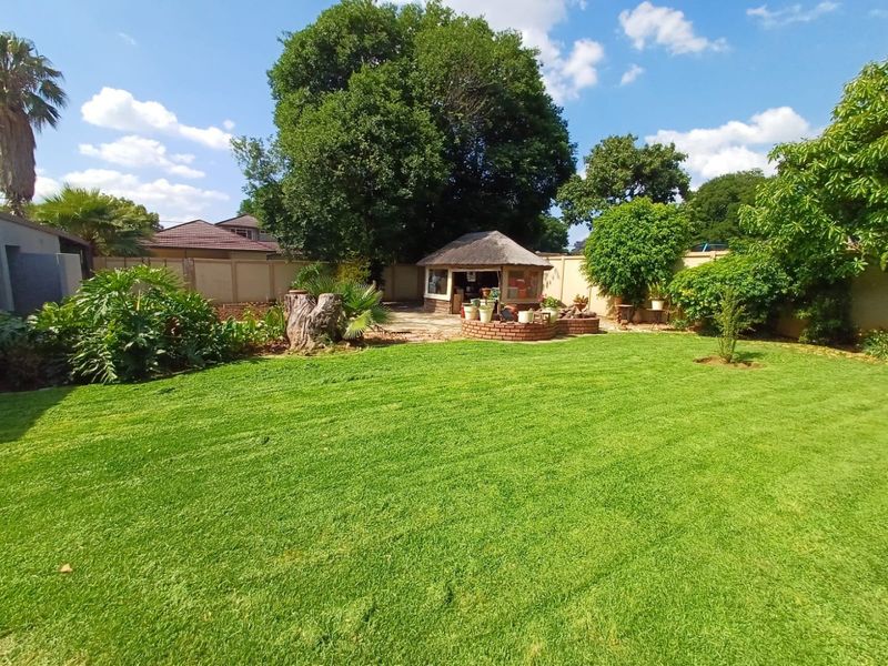 A spacious, well-maintained garden featuring a lush green lawn, mature trees, and a charming thatched lapa area enclosed by a brick boundary wall.