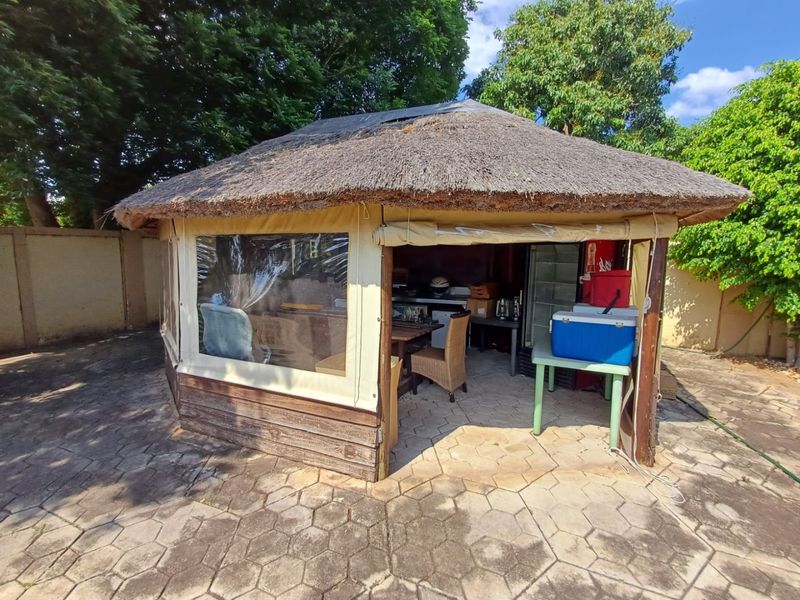 A rustic outdoor entertainment lapa featuring a thatched roof, paved flooring, and roll-up canvas sides, providing a sheltered space for outdoor gatherings.