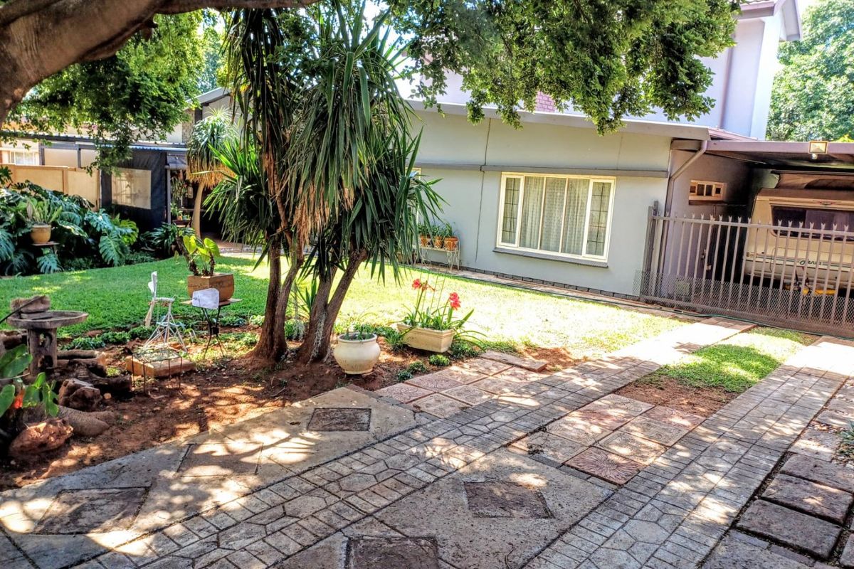 A view of the rear garden area featuring a paved driveway, lawn, mature trees, and a covered carport structure attached to the house.