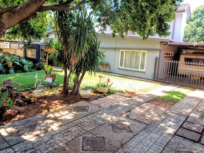 A view of the rear garden area featuring a paved driveway, lawn, mature trees, and a covered carport structure attached to the house.