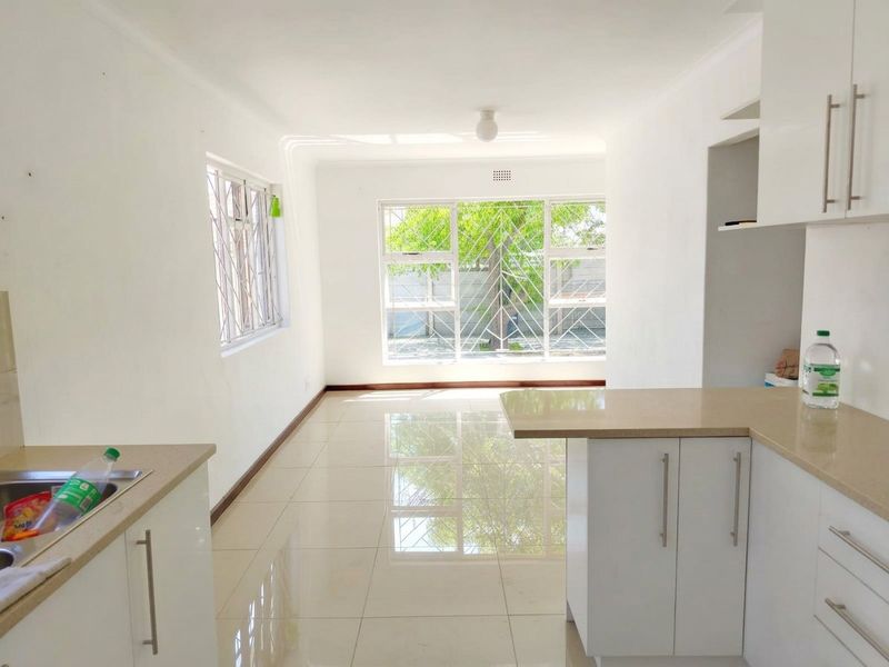 Bright open-plan kitchen and living area featuring modern white cabinetry, stone countertops, tiled flooring, and large windows with security bars.