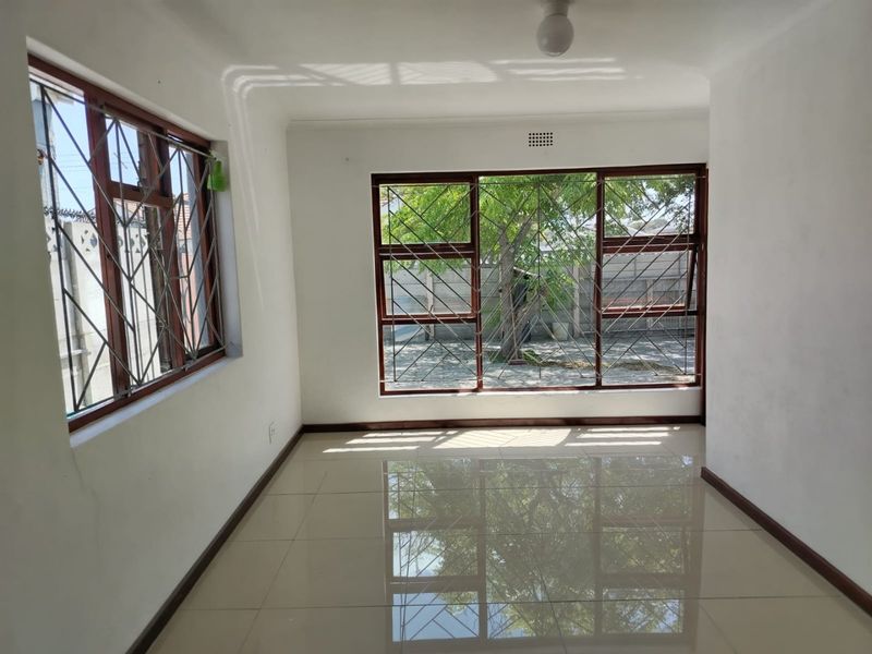 A bright, empty living area featuring polished tiled flooring, timber-framed windows, and security burglar bars, offering a clean canvas for interior design.