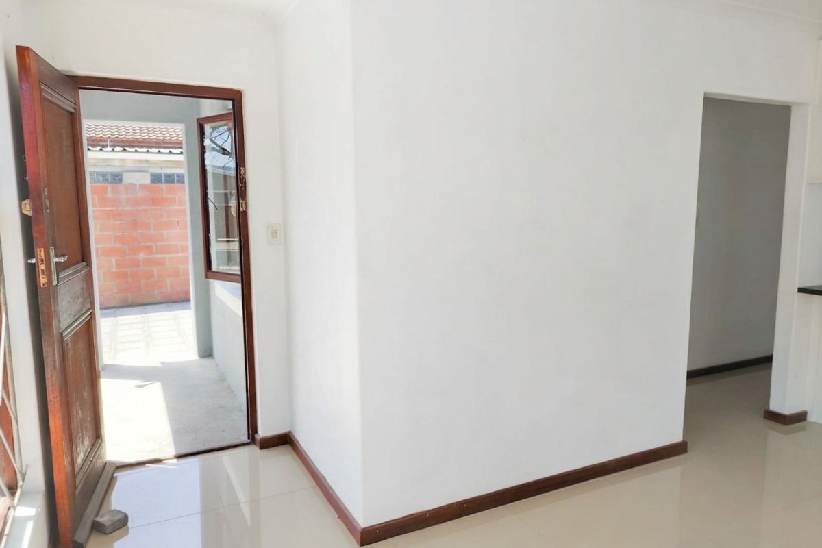 An empty, neutrally painted living area featuring tiled flooring, timber skirting boards, and a wooden door leading to an outdoor space.